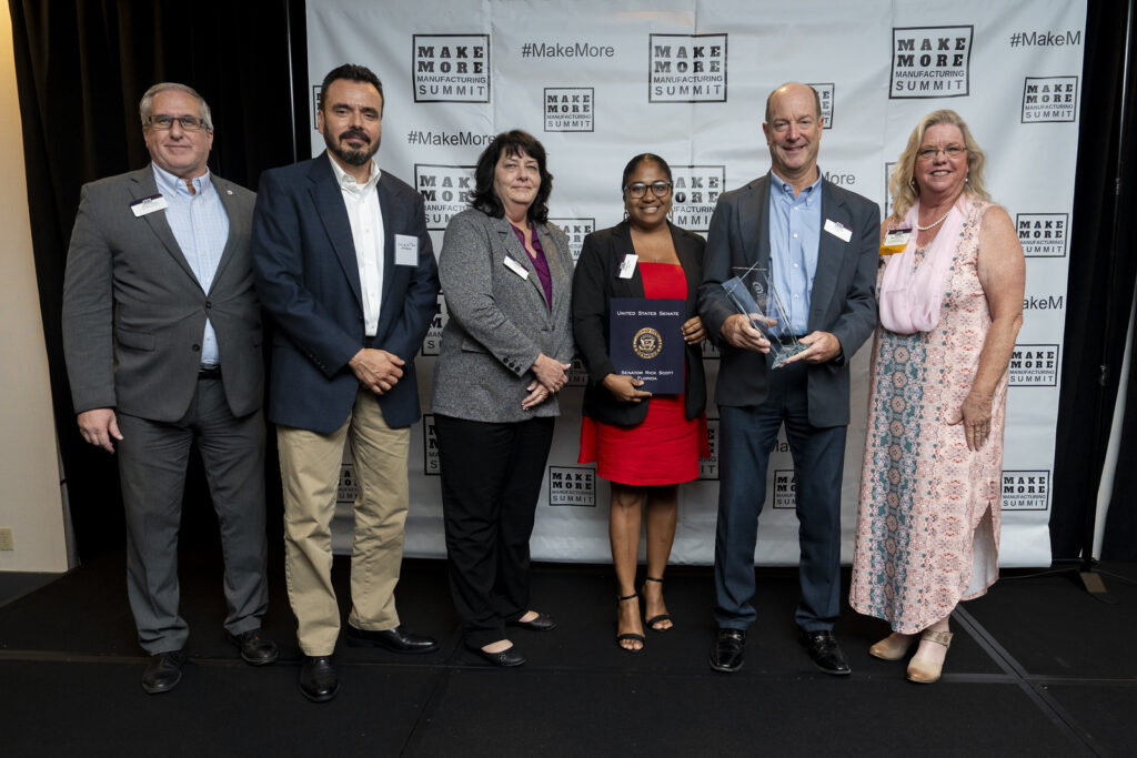 Group photo at a Make More Manufacturing Summit event, seven adults in business attire posing with awards on a stage backdrop.