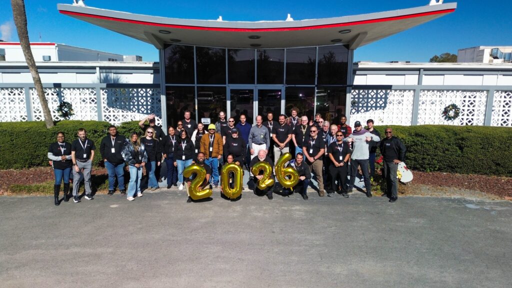 Group of people posing outside a modern building, holding gold balloons that spell '2026' for a team event photo.