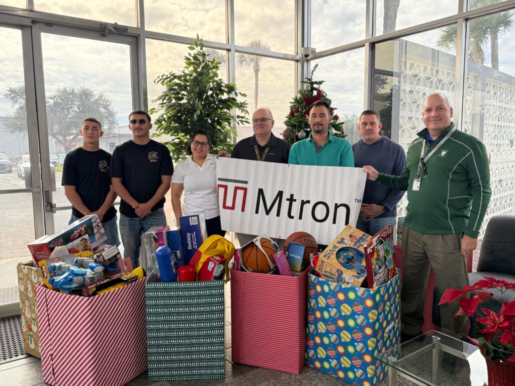 Group of people posing with a banner that reads 'Mtron' behind a row of large gift bags filled with toys and a Christmas tree in the background, indicating a holiday toy drive.