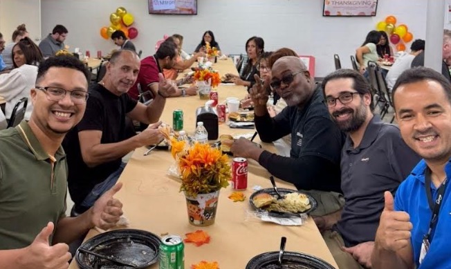 Group of diverse adults seated at a long table sharing a meal, some giving thumbs up, with a fall centerpiece and balloons in the background.
