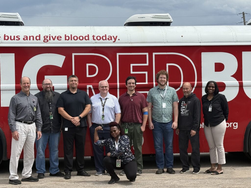 Nine volunteers pose in front of a red blood donation bus with the slogan 'give blood today' visible on the side roped by a large white logo area.