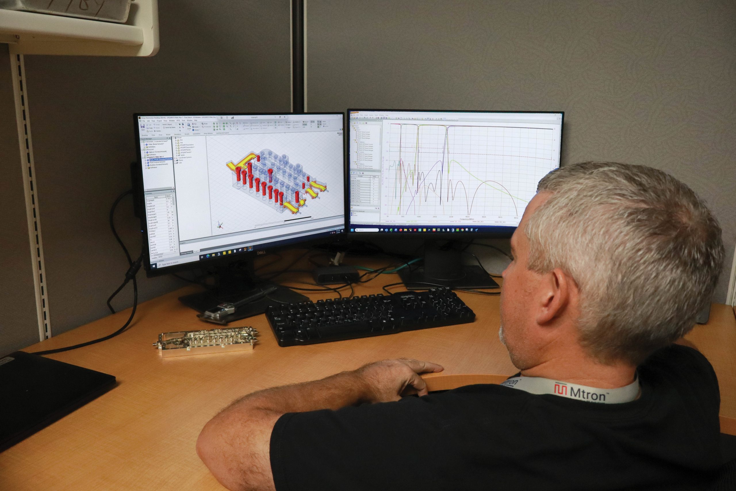 Side profile of a man at a desk working on dual monitors showing a 3D CAD model and data graphs, with a keyboard and electronics on the desk.