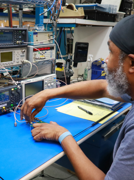 Man with a beard and head wrap works at a blue electronics lab bench, connecting test leads to equipment and instruments on a rack.